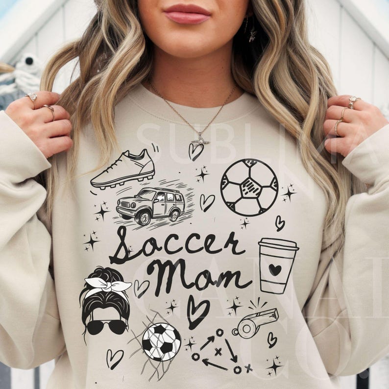 May include: A beige sweatshirt with a black graphic design that says "Soccer Mom" and includes various soccer-related images, including a soccer ball, a soccer cleat, a car, a coffee cup, a whistle, and a soccer net.