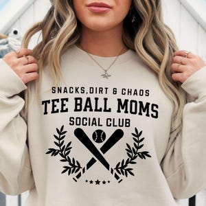 May include: A beige sweatshirt with black text that reads "Snacks, Dirt & Chaos Tee Ball Moms Social Club". The text is surrounded by a graphic of two crossed baseball bats with a baseball and a wreath of leaves.