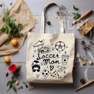 May include: A white canvas tote bag with a black design featuring the text "Soccer Mom" and various soccer-related illustrations, including a soccer ball, cleats, a car, a coffee cup, and a whistle.