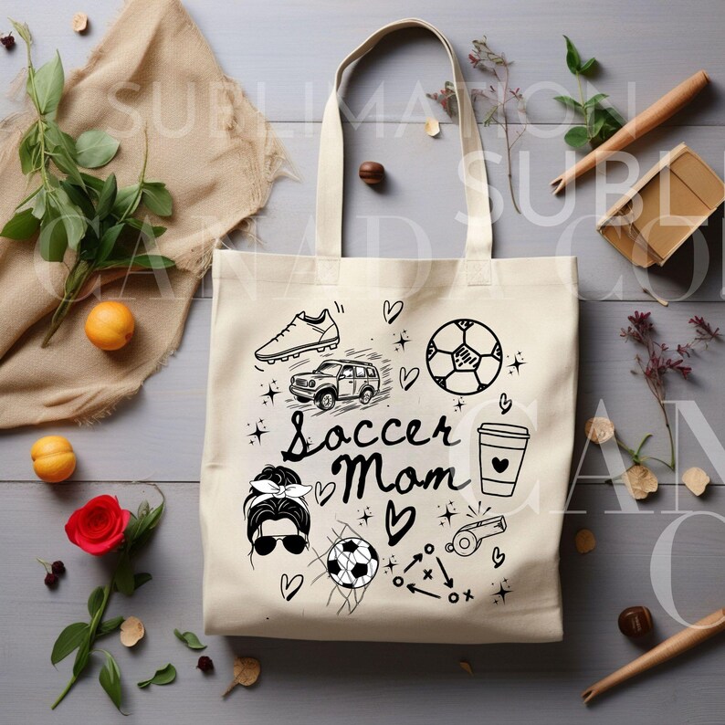 May include: A white canvas tote bag with a black design featuring the text "Soccer Mom" and various soccer-related illustrations, including a soccer ball, cleats, a car, a coffee cup, and a whistle.