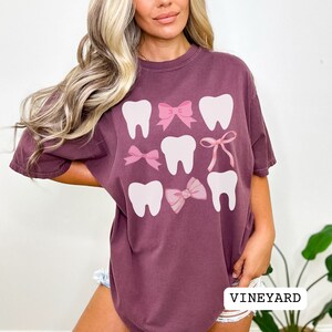 May include: A maroon oversized t-shirt with a graphic design of white teeth and pink bows. The text "VINEYARD" is visible at the bottom of the image.