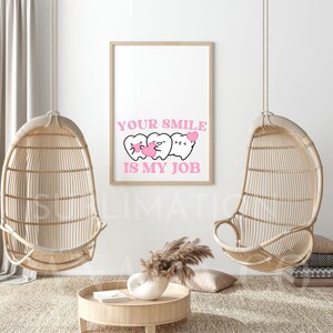 May include: A white framed print with a pink background and the text "YOUR SMILE IS MY JOB". Three cartoon teeth with pink hearts are shown above the text. The print is hanging on a white wall with two wicker hanging chairs on either side.