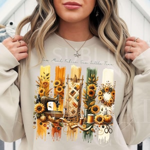 Può includere: Felpa beige con un design a tema cucito. Il disegno include una macchina da cucire vintage, girasoli, una trapunta e il testo "Sew Much Fabric, Sew Little Time". L'opera d'arte utilizza giallo, arancione, verde e marrone.