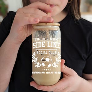 Puede incluir: Un tarro de vidrio transparente con una tapa de bamb&uacute; y una pajita. El tarro est&aacute; lleno de un l&iacute;quido marr&oacute;n y tiene el texto "Soccer Mom Side Line Social Club" y "Warning: May yell at the ref" impreso en &eacute;l.