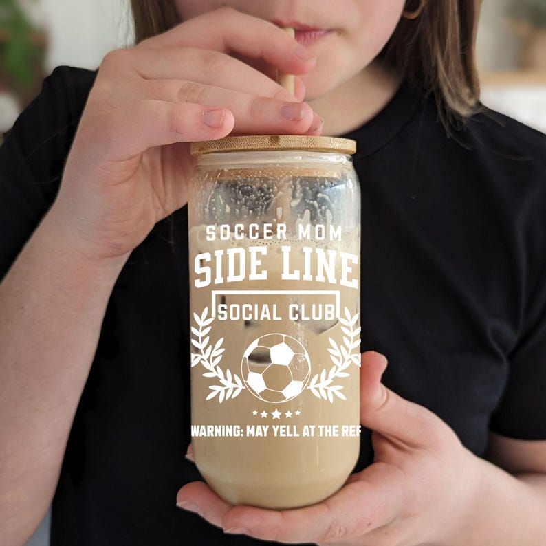 Puede incluir: Un tarro de vidrio transparente con una tapa de bamb&uacute; y una pajita. El tarro est&aacute; lleno de un l&iacute;quido marr&oacute;n y tiene el texto "Soccer Mom Side Line Social Club" y "Warning: May yell at the ref" impreso en &eacute;l.