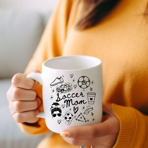May include: White ceramic coffee mug with a black and white illustration of soccer-related items, including a soccer ball, cleats, a car, and a coffee cup. The text "Soccer Mom" is written in black cursive lettering.