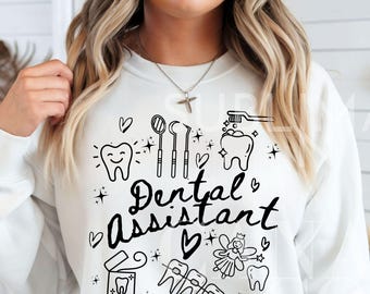 May include: White sweatshirt with black text that reads "Dental Assistant" and a variety of dental-themed illustrations including a toothbrush, a mirror, a tooth, braces, and a fairy.