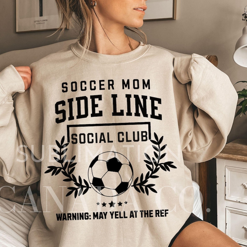 Puede incluir: Sudadera beige con texto negro que dice "Soccer Mom Side Line Social Club" y un gr&aacute;fico de bal&oacute;n de f&uacute;tbol en blanco y negro. El texto "Warning: May yell at the ref" tambi&eacute;n est&aacute; impreso en negro.