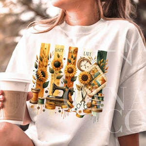 Può includere: Maglietta bianca con un design grafico che presenta girasoli, una macchina da cucire, forbici, bobine di filo e un blocco di trapunta. Il testo "Crazy Quiet Lady" è stampato sul grafico.