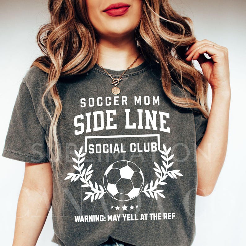 Puede incluir: Una camiseta gris oscura con texto blanco que dice "Soccer Mom Side Line Social Club" con un gr&aacute;fico de bal&oacute;n de f&uacute;tbol y el texto "Warning: May yell at the ref" debajo.