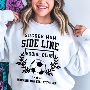 Puede incluir: Sudadera blanca con texto negro que dice "Soccer Mom Side Line Social Club" con un bal&oacute;n de f&uacute;tbol y una corona de laurel. El texto "Warning: May yell at the ref" est&aacute; debajo del dise&ntilde;o.