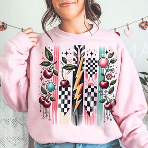 May include: Pink sweatshirt with a graphic design featuring cherries, lightning bolts, and checkered patterns. The design is in shades of red, black, white, and green.