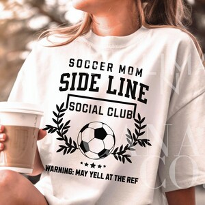Puede incluir: Camiseta blanca con texto negro que dice "Soccer Mom Side Line Social Club" y un gr&aacute;fico de bal&oacute;n de f&uacute;tbol. El texto debajo del gr&aacute;fico dice "Advertencia: Puede gritarle al &aacute;rbitro."