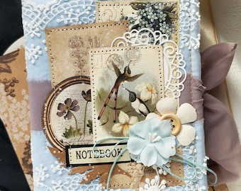 Puede incluir: Un cuaderno cubierto de tela azul claro decorado con encaje blanco, una cinta malva y elementos de papel superpuestos con colibríes, ilustraciones florales y una etiqueta con la palabra NOTEBOOK impresa en tinta negra.