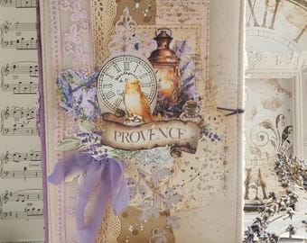 Puede incluir: Un diario rectangular cubierto de tela con un collage de un reloj, un pájaro, una linterna y una pancarta con el texto PROVENCE. La cubierta está decorada con encaje, cinta morada y motivos de lavanda en tonos crema, beige y morado suave.