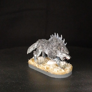 May include: A miniature figurine of a wolf in a running pose. The wolf is painted in shades of gray and white, with red accents in its open mouth. It stands on a small, oval base with tan and green details, suggesting a natural environment.