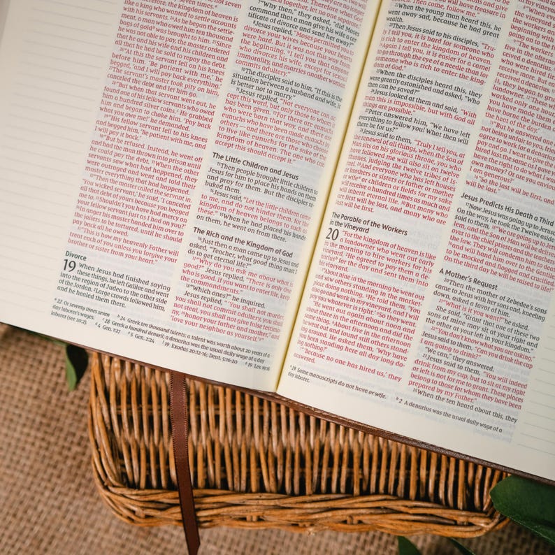 Op de afbeelding: Een open Bijbel in een rieten mand, met een bruine leren bladwijzer. De tekst is in het Engels, met rode hoofdstuknummers. De sc&egrave;ne roept een gevoel van geloof en studie op.
