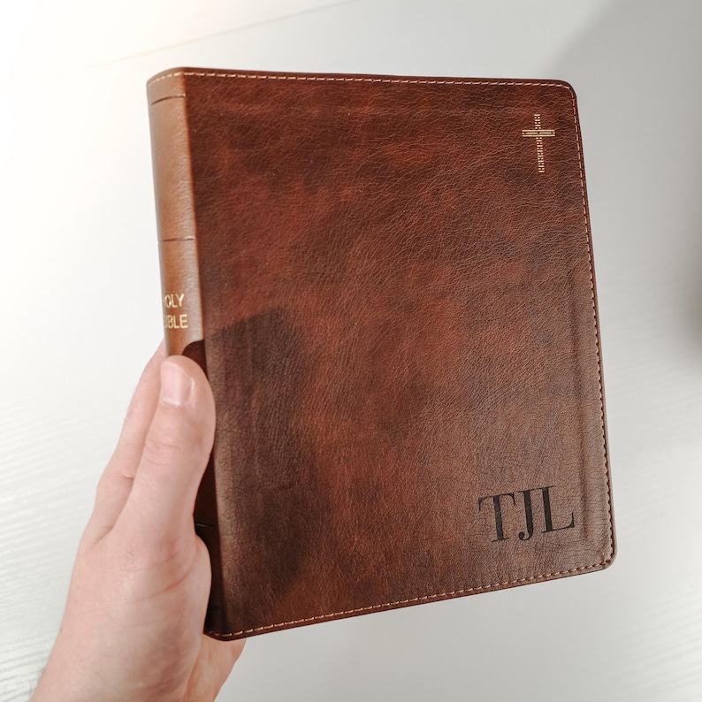 Op de afbeelding: Een bruine leren Bijbel met gouden letters op de rug met de tekst "HOLY BIBLE". De kaft heeft een kruis en de initialen "TJL" gegraveerd op de voorkant. De Bijbel wordt in een hand gehouden.