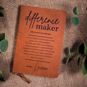 Peut inclure: Un journal en cuir marron avec les mots "difference maker" en écriture noire. La définition de "difference maker" est imprimée en dessous, ainsi que le nom "Lauren". Le journal est sur une surface texturée et beige avec des feuilles vertes.
