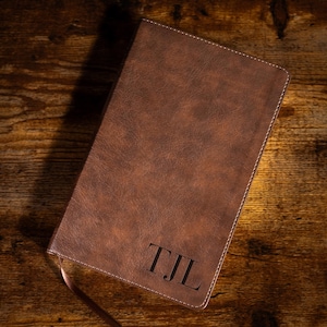 May include: A brown leather journal with the initials "TJL" embossed on the lower right corner. The journal has a textured surface, white stitching along the spine, and a ribbon bookmark. The journal is lying on a wooden surface.
