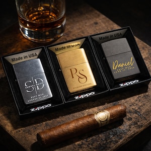Puede incluir: Tres encendedores Zippo grabados en cajas negras, cada uno con iniciales y nombres. Uno es plateado con "SD" y "Samuel Davis", otro es dorado con "RS" y "Richard Stevens", y el tercero es negro con "Daniel". Un cigarro descansa sobre una superficie de madera.