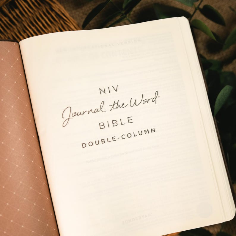 Op de afbeelding: Open Bijbel met de tekst "NIV Journal the Word Bible DOUBLE-COLUMN" in zwart script en blokletters. De pagina's zijn gebroken wit. De achtergrond is een mix van bruin en groen.