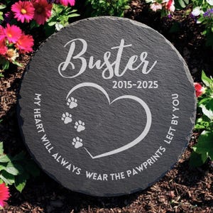 May include: A black slate stone memorial with the name "Buster" and the dates "2015-2025" engraved on it. The stone also features a heart with paw prints and the text "My heart will always wear the pawprints left by you."