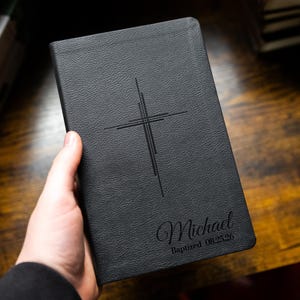 Op de afbeelding: Zwart leren boek met een kruisontwerp en de naam "Michael" en de datum "08.25.26" in reliëf op de kaft. Het boek wordt in een hand gehouden en ligt op een houten oppervlak.