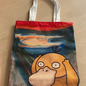 May include: A rectangular fabric tote bag with white handles featuring a printed graphic of a yellow Psyduck character in the style of The Scream painting, set against a dark blue and red swirling background with a landscape scene at the top.