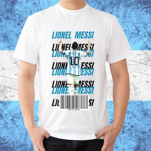 Puede incluir: Camiseta blanca con un gráfico de Lionel Messi con la camiseta de Argentina, con el nombre "Lionel Messi" en texto azul y negro. La camiseta incluye el texto "Made in Argentina" en la parte inferior.