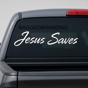 May include: A white decal with the words "Jesus Saves" in a cursive font is displayed on the rear window of a vehicle. The text "BAGEDBRANDING" is above the main text. The vehicle's rear window is visible.