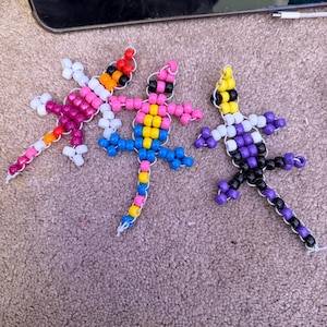 May include: Three lizard-shaped figures crafted from colorful plastic pony beads strung on white cord. The left lizard features white, pink, and orange beads. The center lizard uses pink, yellow, and blue beads. The right lizard has yellow, white, purple, and black beads.