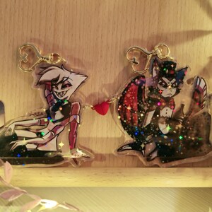 May include: Two glitter-filled acrylic charms featuring cartoon characters. The first charm is a red and white character with a devilish grin. The second charm is a black and white character with a cat-like face and a top hat. Both charms are attached to gold keychains.