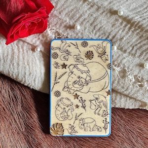 Puede incluir: Una placa de madera con un borde azul presenta un diseño grabado con láser de una madre elefante con su bebé. El diseño también incluye otros elefantes, una cigüeña y flores.