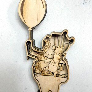 May include: Wooden layered art piece featuring Winnie the Pooh, Piglet, and Eeyore. Pooh holds a balloon. The artwork is made of light-colored wood and has a three-dimensional effect, with detailed cutouts.