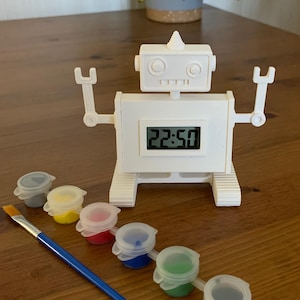 May include: A white robot-shaped digital clock showing 22:50. The clock has arms and a head with circular details. Beside it are small paint pots in various colours and a blue-handled paintbrush, all on a wooden surface.