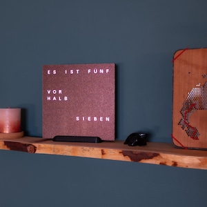 Könnte beinhalten: Ein Holzregal mit einer rot-braunen Uhr, auf der "Es ist fünf vor halb sieben" steht, und einer Holz-Wanddekoration mit einem roten Schnurmuster.