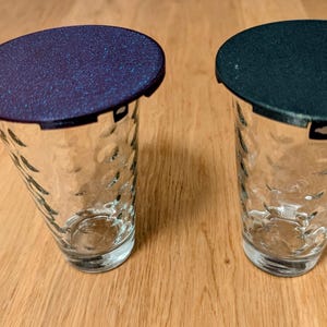 Könnte beinhalten: Zwei klare Glasbecher mit strukturierten Seiten, jeweils mit einem Deckel. Ein Deckel ist dunkelblau mit einer gesprenkelten Oberfläche, der andere Deckel ist dunkelgrün.