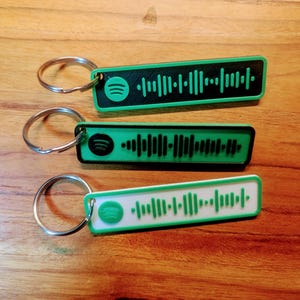 Op de afbeelding: Drie sleutelhangers met zwarte, witte en groene rechthoekige labels met een Spotify-logo en een barcode-achtig patroon. Elke sleutelhanger heeft een zilveren metalen ring.