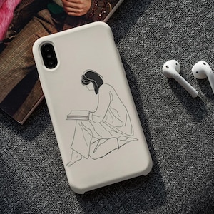 May include: A white phone case featuring a black line drawing of a person reading a book. Two white wireless earbuds are beside the phone case. The items are on a grey textured surface.
