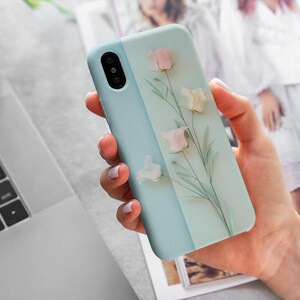 May include: A pale blue phone case with a floral design. The case has a two-tone design with a lighter and darker blue section. The floral design includes pink and white flowers with green stems.