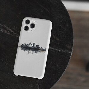 May include: A white smartphone case with a black and grey skyline design and a mirrored reflection. The case has a glossy finish and a camera module with three lenses. The case is on a dark, textured surface.