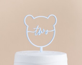 May include: Light blue cake topper shaped like a bear with the word "two" inside. The topper is on top of a white frosted cake.