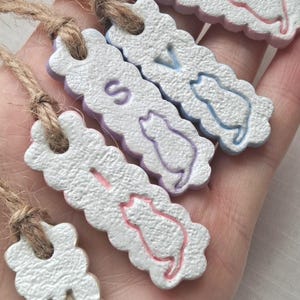 Puede incluir: Cinco etiquetas blancas con bordes festoneados con siluetas de gatos y letras, suspendidas por hilo. Las etiquetas son de varios colores pastel: rosa, azul y morado. Las letras deletrean "M", "R", "S" y "V".