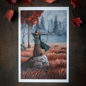 May include: A print depicting a woman archer in a forest. She is poised on a rock, drawing back a bow. The artwork uses greens, browns, and reds, with trees and a misty sky in the background. Red autumn leaves are scattered around the print.