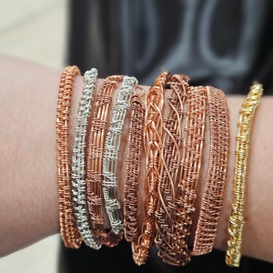 May include: A stack of handmade wire bracelets in various shades of copper and silver. The bracelets are woven with a variety of patterns, including a simple braid and a more intricate basket weave.