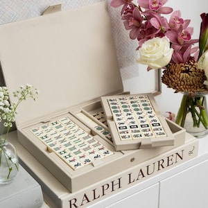 Puede incluir: Un juego de Mahjong de cuero sint&eacute;tico beige con tapa abierta, que revela fichas con s&iacute;mbolos coloridos. El juego est&aacute; sobre una pila de libros, uno de ellos con la etiqueta "RALPH LAUREN". Un jarr&oacute;n de flores, incluyendo orqu&iacute;deas y rosas, est&aacute; a la derecha.