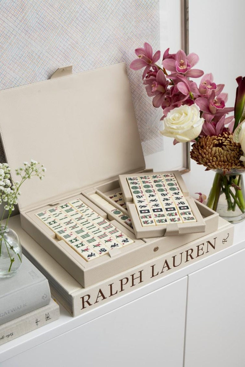 Puede incluir: Un juego de Mahjong de cuero sint&eacute;tico beige con tapa abierta, que revela fichas con s&iacute;mbolos coloridos. El juego est&aacute; sobre una pila de libros, uno de ellos con la etiqueta "RALPH LAUREN". Un jarr&oacute;n de flores, incluyendo orqu&iacute;deas y rosas, est&aacute; a la derecha.