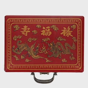 May include: A red rectangular box with a gold border and handle. The box features gold illustrations of a phoenix and a dragon, along with Chinese characters and cloud motifs. The overall design is ornate and traditional.