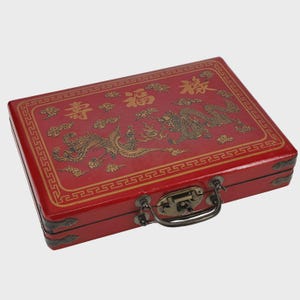 May include: A red rectangular box with gold Chinese characters and illustrations of a dragon and phoenix. The box has a metal clasp and handle, and a decorative border.